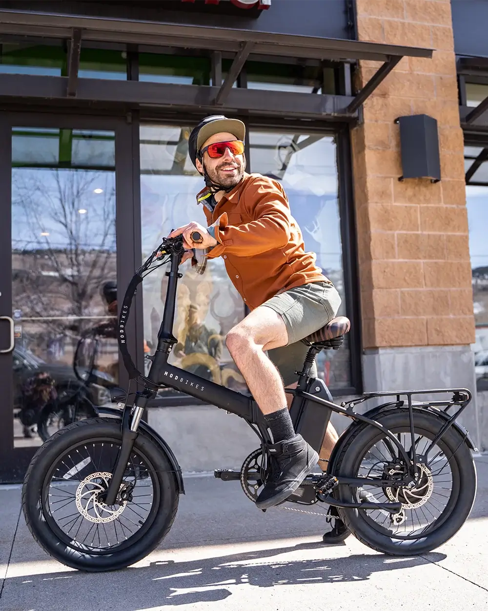 Man riding the MOD City+ 3 Foldable Electric Bike outside a store, showcasing its sleek design and full-suspension features.